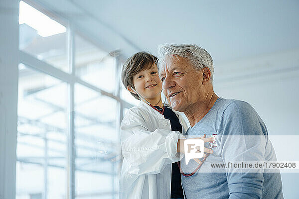 Smiling boy imitating as doctor checking grandfather with stethoscope at home