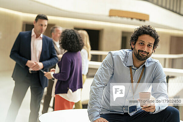 Smiling businessman holding coffee cup with colleagues discussing in background