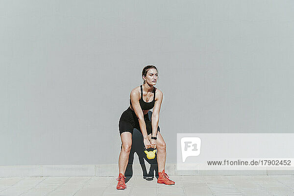 Young woman with kettlebell exercising in front of gray wall