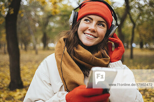 Lächelnde junge Frau hält ihr Mobiltelefon in der Hand und hört im Park Musik über Kopfhörer
