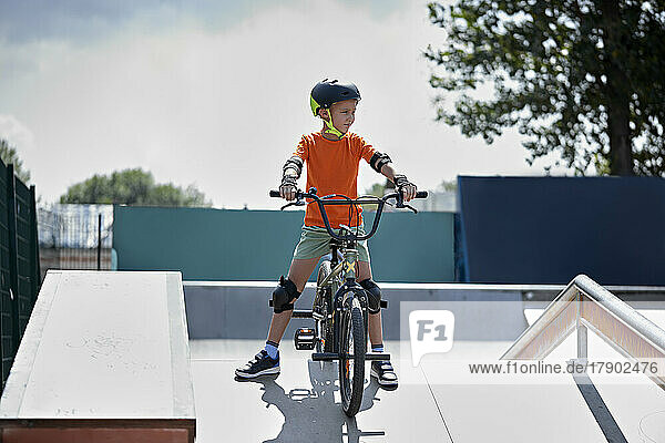 Boy with protective gears sitting on BMX bike at skateboard park