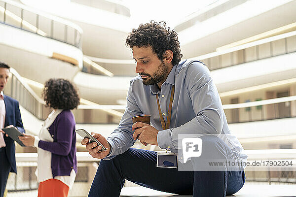 Businessman text messaging on smart phone with colleagues discussing in background at office
