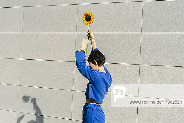 Frau mit erhobenen Armen hält an einem sonnigen Tag eine Sonnenblume an der Wand