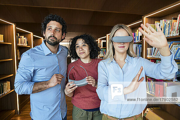 Woman with VR glasses gesturing by colleagues in illuminated library