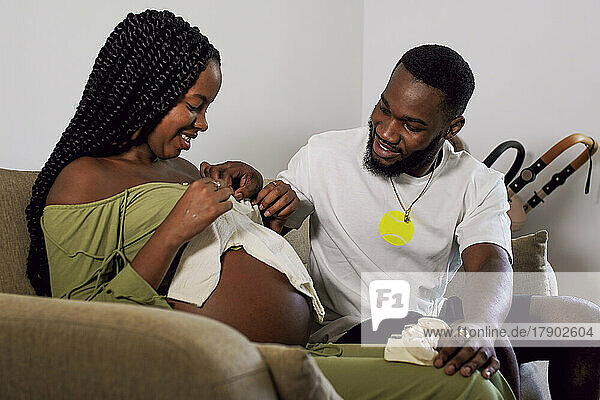 Mann schaut sich Babykleidung an  die eine schwangere Frau zu Hause hält