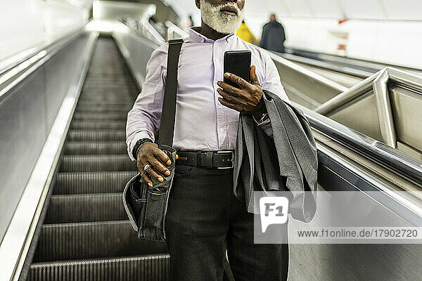 Senior businessman with suit and mobile phone carrying laptop bag on escalator