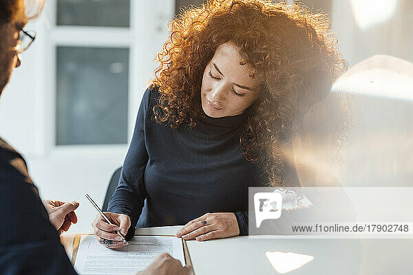 Businesswoman with curly hair signing document sitting at desk in office