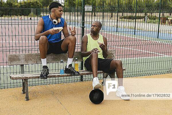 Father and son eating sandwiches sitting on bench at basketball court