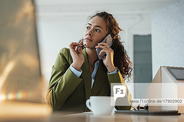 Thoughtful businesswoman talking on smart phone sitting by model house at desk in office