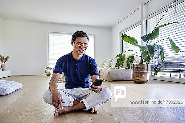 Lächelnder reifer Mann mit Smartphone  der zu Hause auf dem Boden sitzt