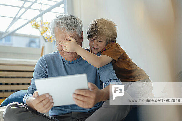 Boy covering eyes of grandfather with tablet PC at home