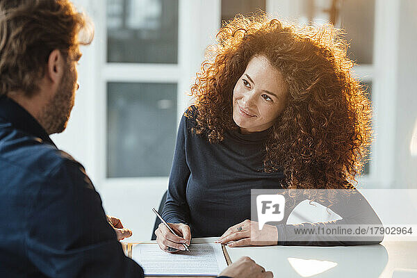 Smiling young businesswoman signing contract and looking at businessman in office