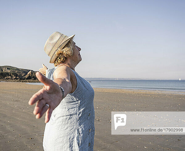 Ältere Frau mit ausgestreckten Armen steht an einem sonnigen Tag am Strand