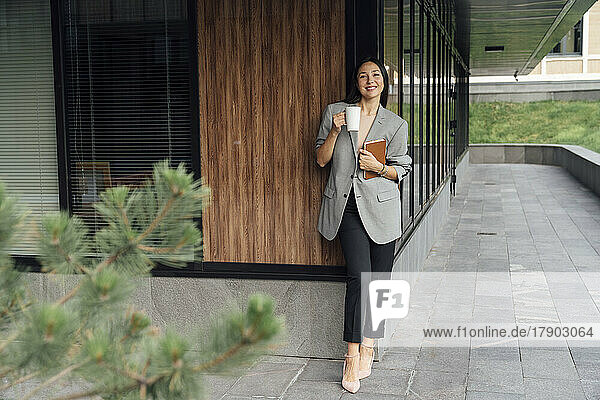 Smiling businesswoman with coffee mug and book standing outside office building