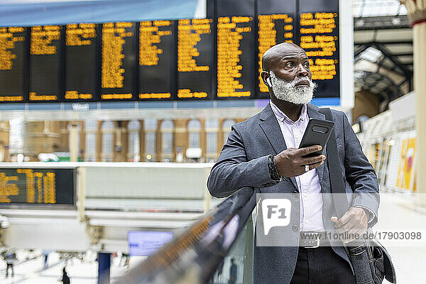 Businessman with smart phone listening to music on wireless in-ear headphones at station