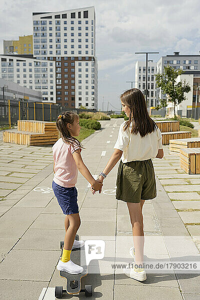 Girl holding hand of sister teaching longboard skating at footpath on sunny day