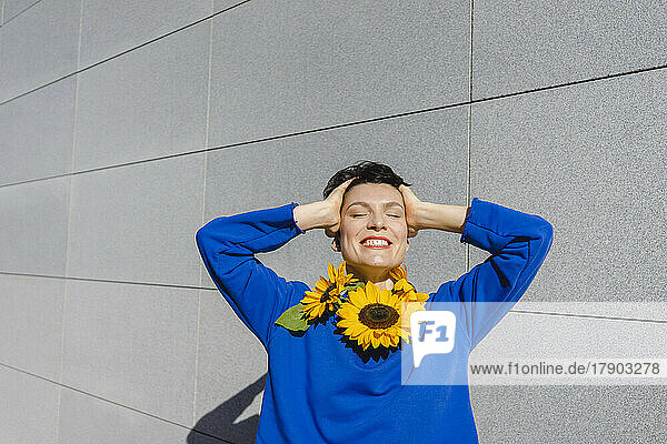 Happy woman with sunflowers around neck in front of wall on sunny day
