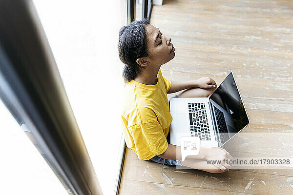 Junge Frau sitzt mit geschlossenen Augen auf dem Boden  lehnt an einem Fenster und hat einen Computer auf dem Schoß