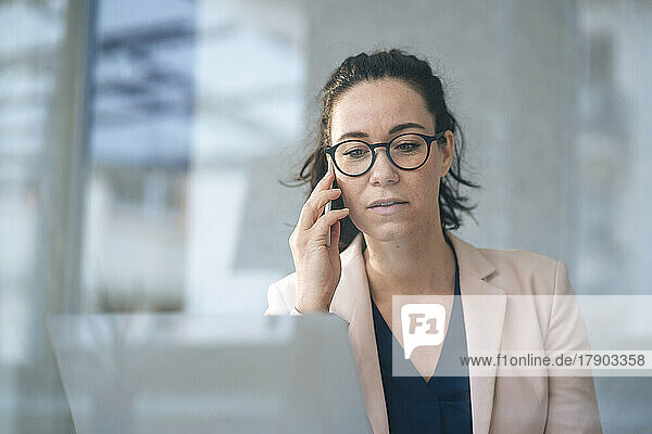 Businesswoman talking on mobile phone sitting with laptop in front of gray wall seen through glass