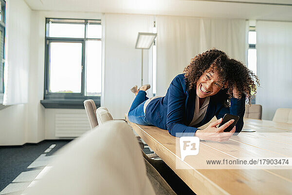 Fröhliche Geschäftsfrau mit Smartphone am Konferenztisch im Büro liegend