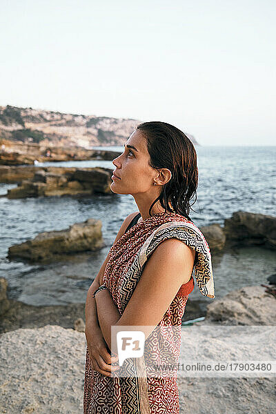 Thoughtful woman with arms crossed wrapped in blanket by sea