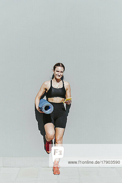 Smiling woman with mobile phone and exercise mat leaning on gray wall