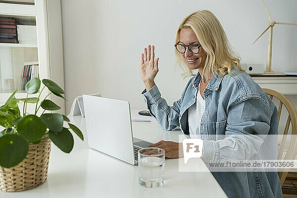 Smiling freelancer waving hand on video call through laptop at home