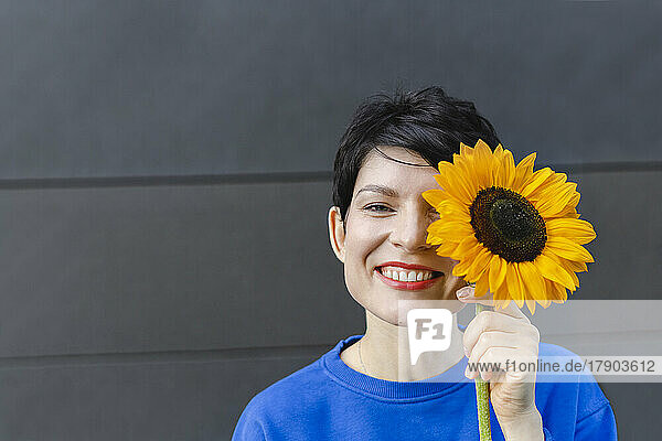 Happy woman holding sunflower over face standing in front of wall