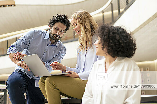 Happy blond businesswoman discussing over laptop with colleagues in corridor