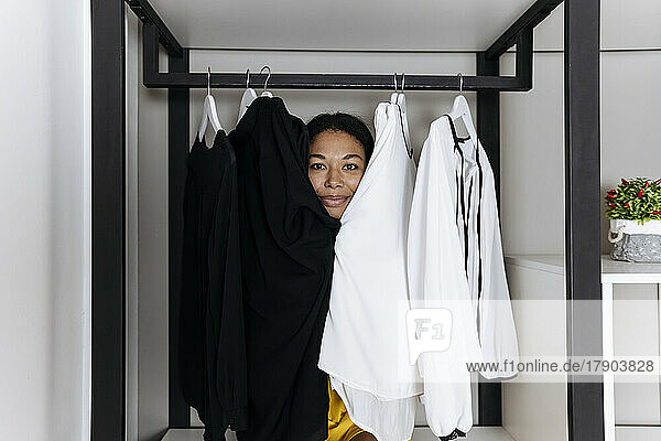 Woman sitting inside closet and peeking out from behind clothes