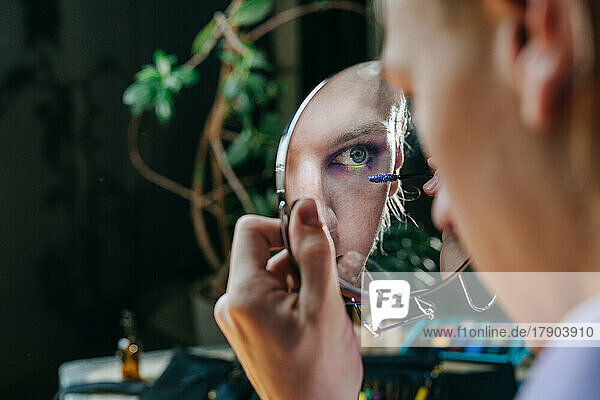 Reflection of man applying mascara in hand mirror