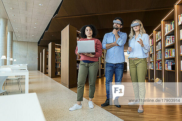 Mann und Frau schauen durch eine VR-Brille einem Kollegen zu  der einen Laptop in der Bibliothek hält