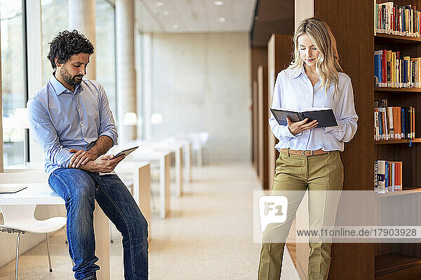 Professor using tablet PC by colleague reading book in library