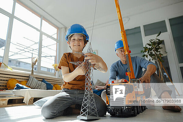 Smiling boy imitating as engineer holding electricity pylon model with grandfather in background at home