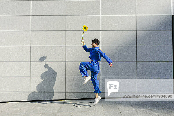 Frau mit Sonnenblume springt an sonnigem Tag vor Mauer