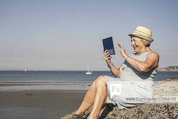Happy senior woman waving hand on video call sitting at beach