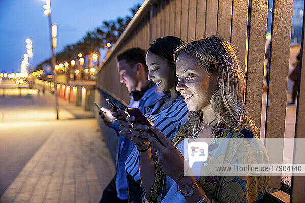 Smiling women and man text messaging through mobile phones in front of railing at night
