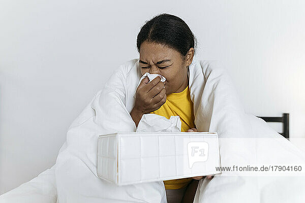 Sick woman sitting on bed wrapped in a blanket blowing nose in a tissue