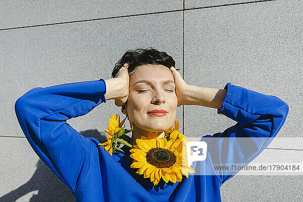 Smiling woman with sunflowers around neck standing in front of wall on sunny day