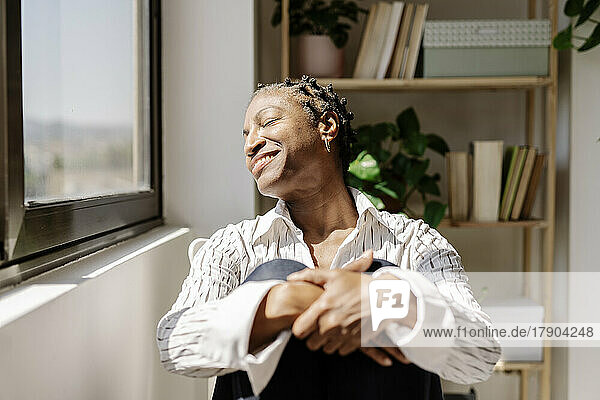 Smiling woman enjoying sunlight sitting by window at home