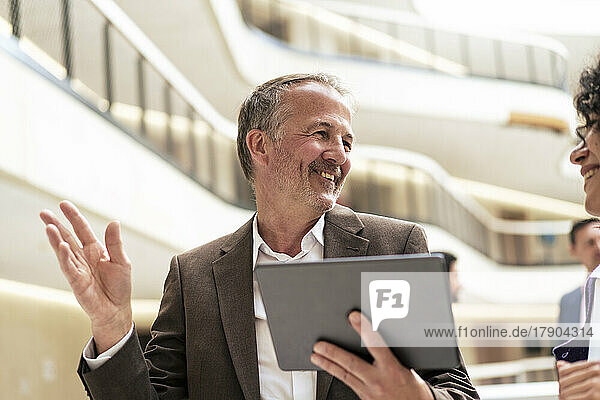 Smiling businessman holding tablet PC discussing with colleague in corridor