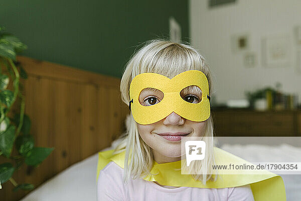 Smiling blond girl wearing yellow mask and cape at home