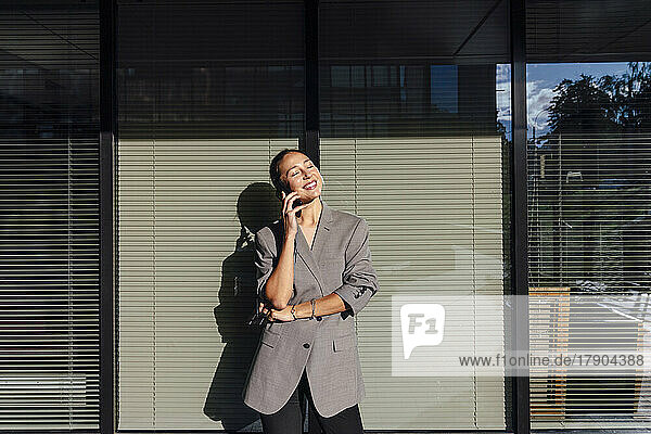 Smiling businesswoman with eyes closed talking on mobile phone in front of glass wall