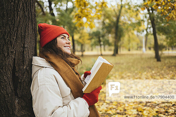 Thoughtful smiling woman with book leaning on tree trunk at park