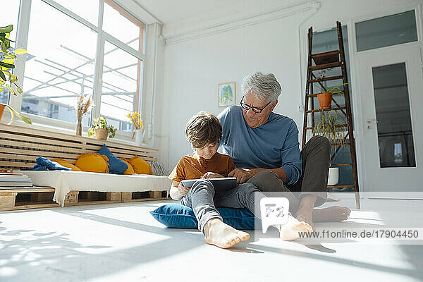 Smiling grandson using tablet computer by grandfather in living room at home