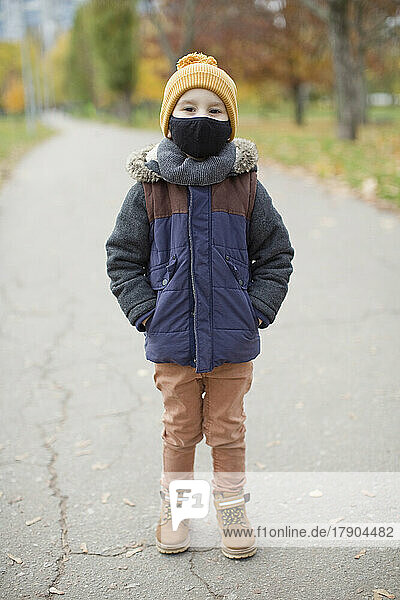 Boy standing with hands in pockets wearing protective face mask on footpath
