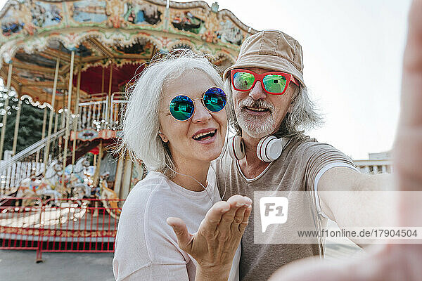 Senior man with mature woman taking selfie in front of carousel at amusement park