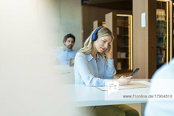 Businesswoman listening music on headphones using smart phone sitting at desk in office