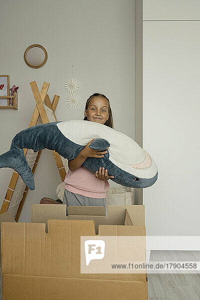 Lächelndes Mädchen mit Hai-Spielzeug  das zu Hause neben der Kiste steht