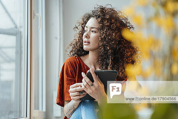 Thoughtful woman with tablet PC sitting by window at home